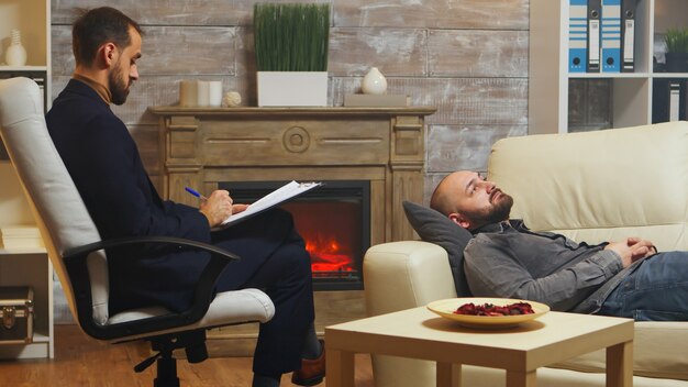 Бородатый мужчина, лежа на диване во время терапии для пар, говорит о конфликтах в отношениях с женой.