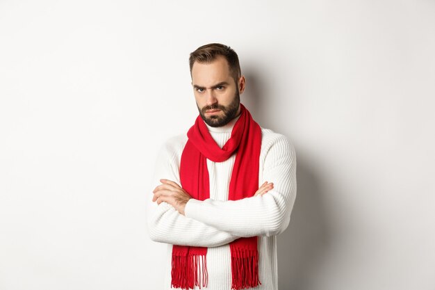Бородатый мужчина выглядит сердитым и обиженным на вас, скрестив руки на груди в оборонительной позе, дуется, стоя в рождественском свитере на белом фоне.