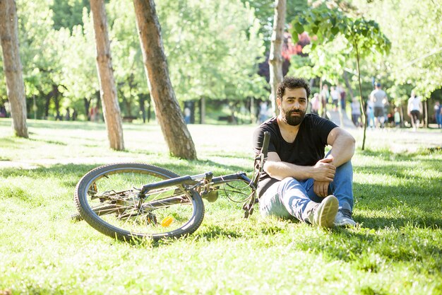 Бородатый мужчина смотрит в камеру рядом со своим велосипедом, сидя на траве в парке