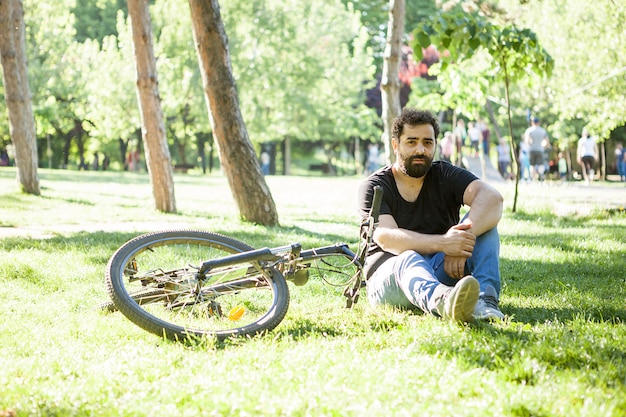 公園の芝生の上に座っている彼の自転車の横にあるカメラを見ているひげを生やした男