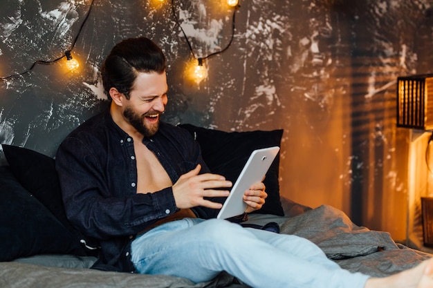 Bearded man in morning time, lying on bed with tablet and smiling at modern badroom.