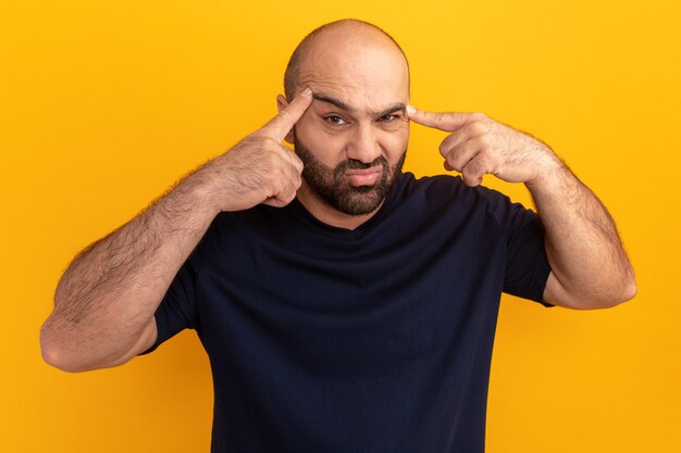 Bearded man in navy t-shirt  being confused pointing with index fingers at his temples standing over orange wall