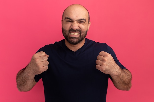 Free photo bearded man in navy t-shirt  clenching fists with annoyed expression standing over pink wall
