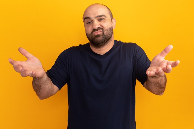 Bearded man in navy t-shirt  confused and displeased with arms raised standing over orange wall