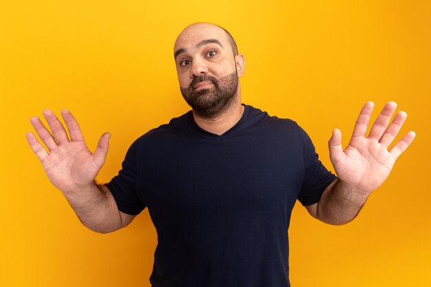 Бородатый мужчина в темно-синей футболке смущенно поднимает руки, не имея ответа, стоит над оранжевой стеной