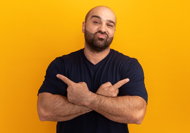 Bearded man in navy t-shirt  crossing hands pointing with index fingers to the sides smiling standing over orange wall