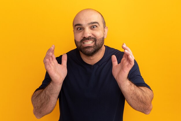 Бородатый мужчина в темно-синей футболке эмоциональный и счастливый с поднятыми руками стоит над оранжевой стеной