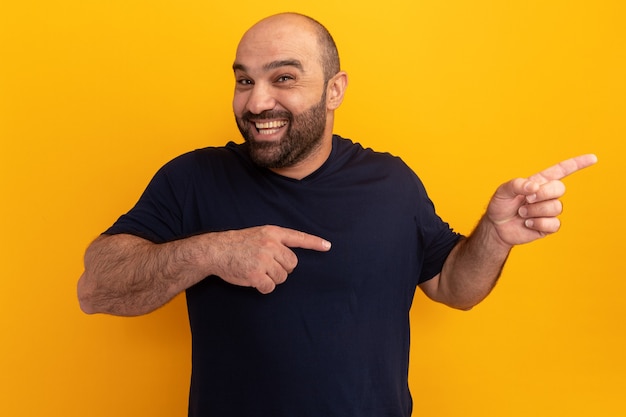 Бородатый мужчина в темно-синей футболке счастливый и веселый улыбается, указывая указательными пальцами в сторону, стоя над оранжевой стеной