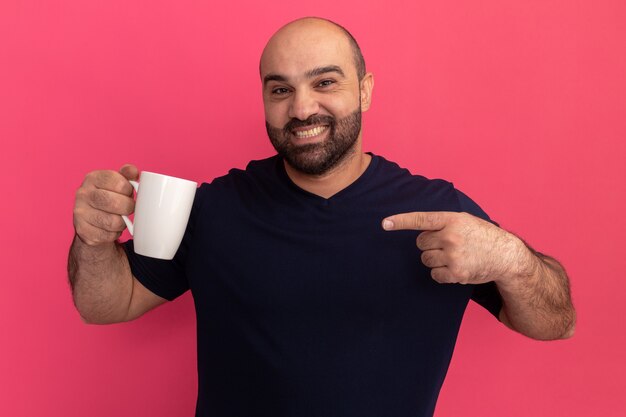 검지 손가락으로 가리키는 컵을 들고 해군 티셔츠에 수염 난 남자가 분홍색 벽 위에 서 행복하고 긍정적 인 미소