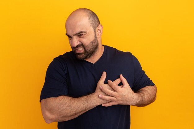Bearded man in navy t-shirt holding hands on chest looking unwell standing over orange wall