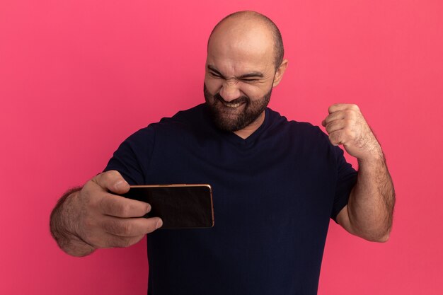 Bearded man in navy t-shirt holding smartphone looking at it happy and excited clenching fist standing over pink wall