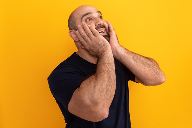 Bearded man in navy t-shirt lookign up amazed and surprised with hands on his face standing over orange wall