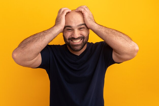 Bearded man in navy t-shirt looking confused and frustrated with hands on his head standing over orange wall