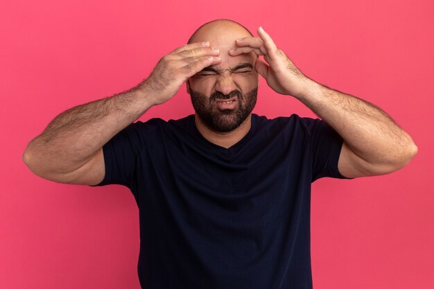 Bearded man in navy t-shirt looking unwell touching his head with annoyed expression suffering from strong headache standing over pink wall