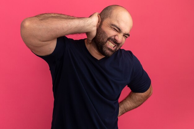 Free photo bearded man in navy t-shirt looking unwell touching his neck feeling pain standing over pink wall