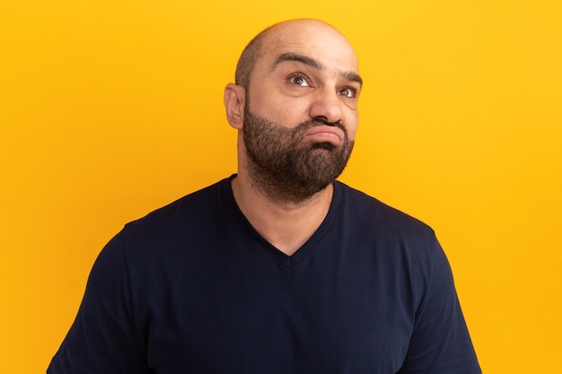 Bearded man in navy t-shirt looking up puzzled standing over orange wall