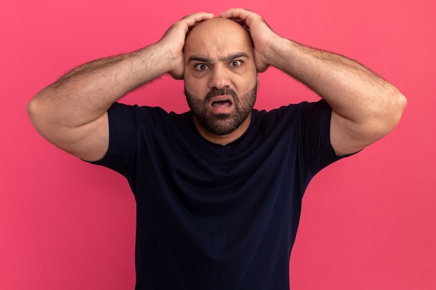 Bearded man in navy t-shirt  mazed and worried with raised hands over head standing over pink wall