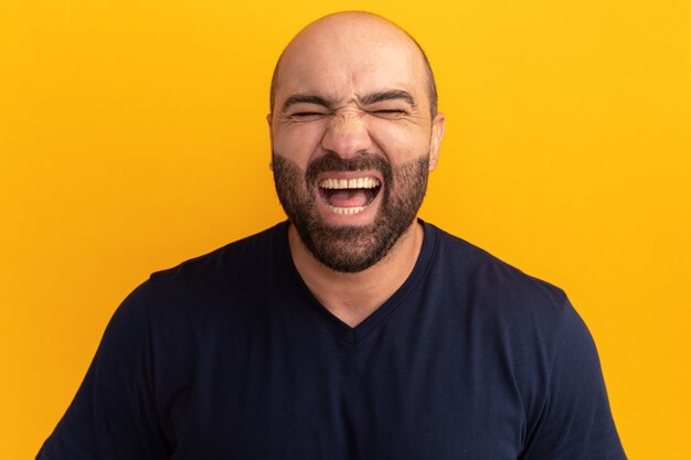 Bearded man in navy t-shirt screaming happy and excited standing over orange wall