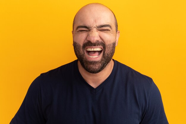 Бородатый мужчина в темно-синей футболке кричит от счастья и возбуждения, стоя у оранжевой стены