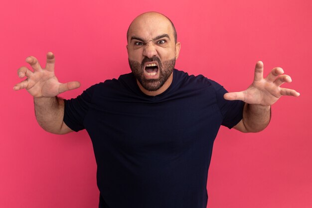 Bearded man in navy t-shirt  shouting with angry expression with raised arms frightening standing over pink wall