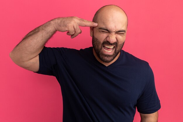 Bearded man in navy t-shirt shouting with annoyed expression pointing with index finger at his temple standing over pink wall