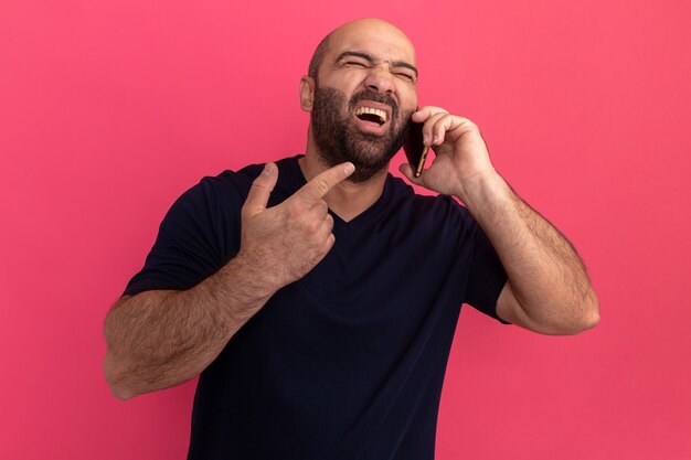 Бородатый мужчина в темно-синей футболке разговаривает по мобильному телефону и кричит от разочарования, стоя у розовой стены