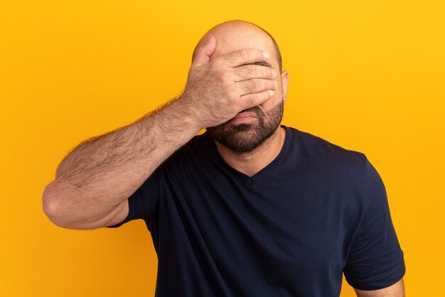 Bearded man in navy t-shirt tired and disappointed covering face with hand standing over orange wall
