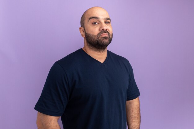 Bearded man in navy t-shirt  wirh serious face standing over purple wall