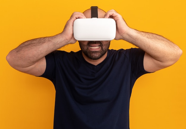 Bearded man in navy t-shirt with glasses of virtual reality looking amazed and surprised standing over orange wall