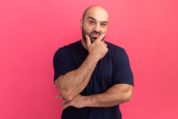 Bearded man in navy t-shirt  with hand on chin thinking standing over pink wall