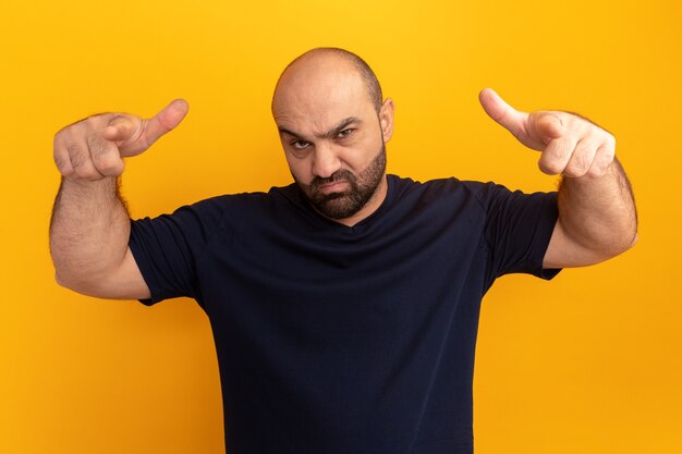 Bearded man in navy t-shirt  with serious face pointing with index fingers  standing over orange wall