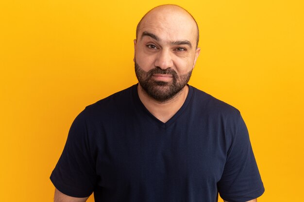 Bearded man in navy t-shirt  with serious face standing over orange wall