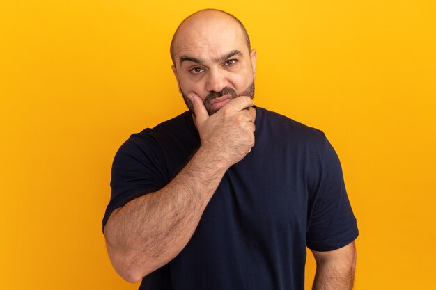 Bearded man in navy t-shirt  with skeptic expression with nad on chin standing over orange wall