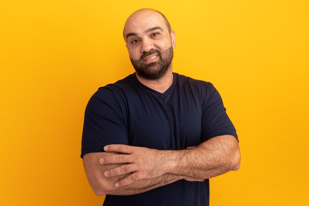 Bearded man in navy t-shirt  with skeptic smile on face with arms crossed standing over orange wall