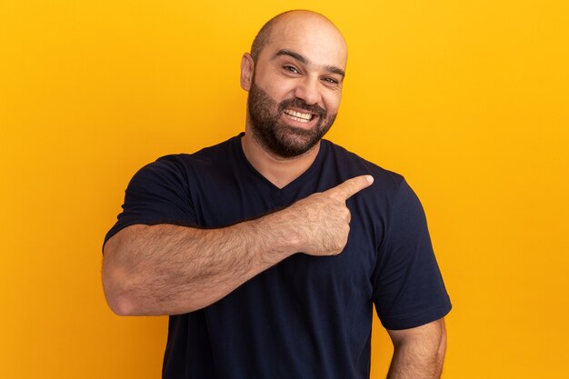 Bearded man in navy t-shirt  with smile on face pointing with index finger to the side standing over orange wall