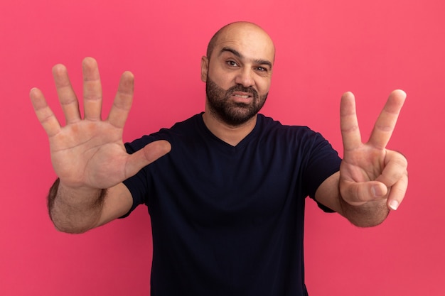 Bearded man in navy t-shirt  with smile on face showing number seven standing over pink wall