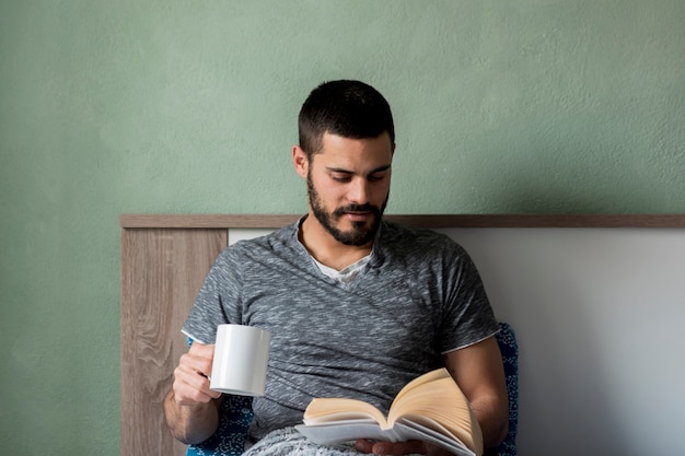 Foto gratuita libro di lettura barbuto dell'uomo e tazza di tenuta