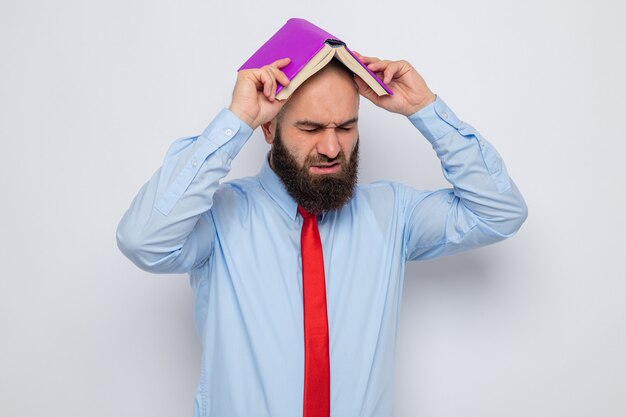 Бородатый мужчина в красном галстуке и синей рубашке держит книгу над головой, выглядит усталым и раздраженным