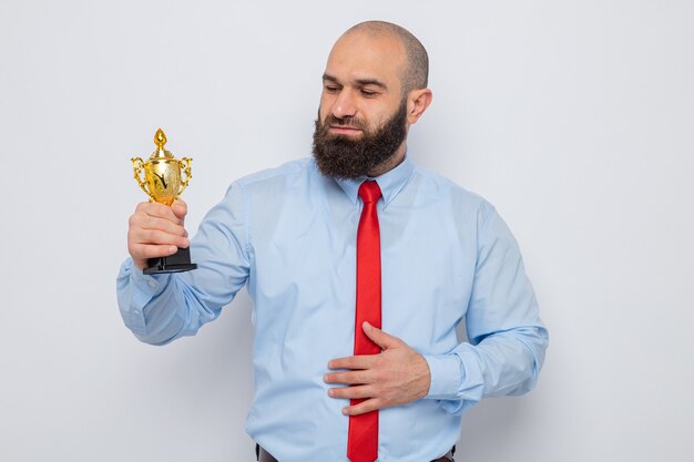 Bearded man in red tie and blue shirt holding trophy looking at it with smile on happy face standing over white background