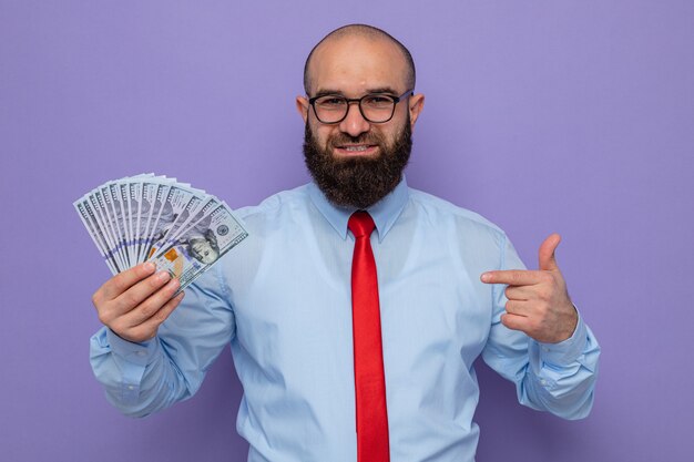 Bearded man in red tie and blue shirt wearing glasses holding cash pointing with index finger at money looking at camera smiling cheerfully standing over purple background