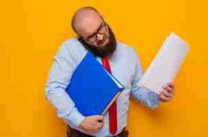 Foto gratuita uomo barbuto in cravatta rossa e camicia blu con gli occhiali che tengono la cartella dell'ufficio e i documenti impegnati e stressati a parlare al cellulare on