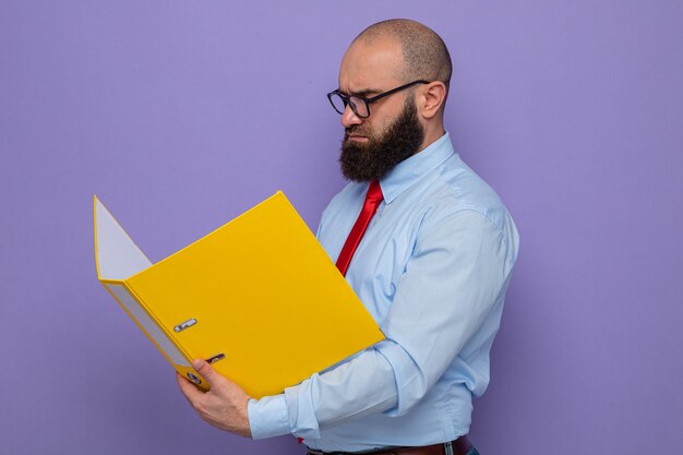 Bearded man in red tie and blue shirt wearing glasses holding office folder looking it with serious face