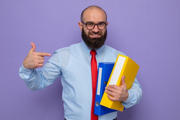 Uomo barbuto in cravatta rossa e camicia blu con gli occhiali che tengono le cartelle dell'ufficio che puntano con il dito indice a guardarli sorridendo allegramente