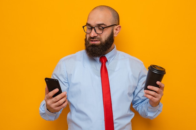 Бородатый мужчина в красном галстуке и синей рубашке в очках держит смартфон и чашку кофе счастливым и позитивным, улыбающимся, уверенно стоящим на оранжевом фоне