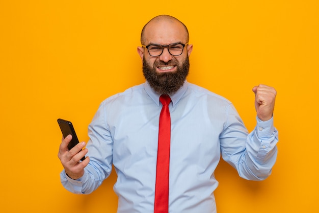 Бородатый мужчина в красном галстуке и синей рубашке в очках держит смартфон счастливым и взволнованным, поднимая кулак, как победитель