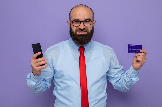 빨간 넥타이와 파란 셔츠를 입은 수염 난 남자는 안경 스마트폰과 신용 카드를 착용하고 카메라를 보고 행복하고 긍정적인 미소를 보라색 배경 위에 밝게 서 있습니다.