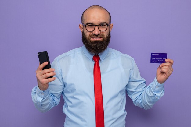 紫色の背景の上に元気に立って幸せで前向きな笑顔のカメラを見てメガネスマートフォンとクレジットカードを身に着けている赤いネクタイと青いシャツのひげを生やした男