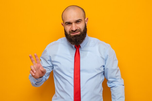 Бородатый мужчина в красном галстуке и рубашке смотрит в камеру, весело улыбаясь, показывая номер два с пальцами, стоящими на оранжевом фоне