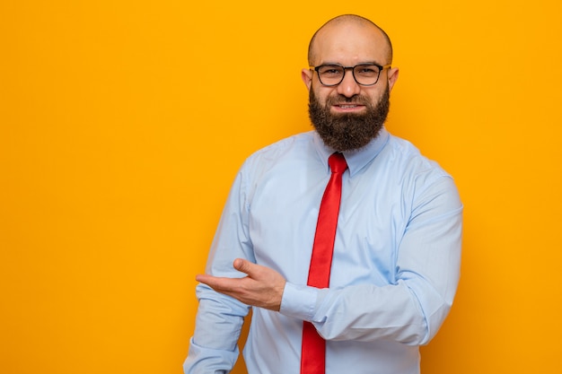 Bearded man in red tie and shirt wearing glasses looking at camera smiling presenting copy space with arm of his handstanding over orange background