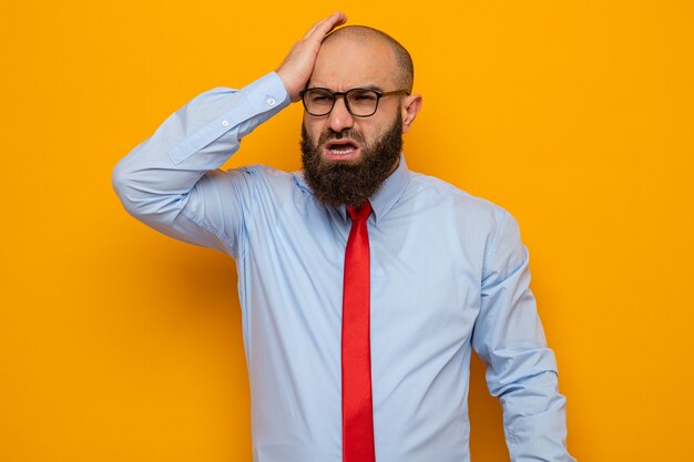 Bearded man in red tie and shirt wearing glasses looking confused holding hand on his head for mistake standing over orange background